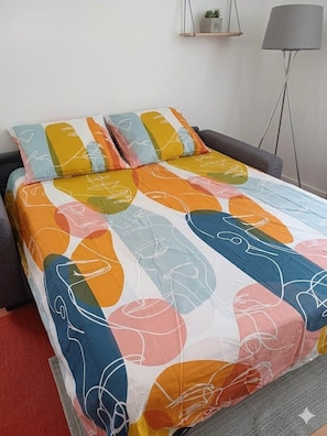 Una cama con una colcha y almohadas de diseño colorido, una lámpara de pie gris y una planta pequeña en un estante.