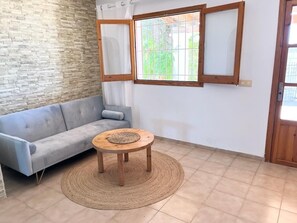 Ein Zimmer mit grauem Sofa, einem runden Holztisch mit geflochtener Unterlage, einem Fenster mit Holzrahmen und einem Fliesenboden.