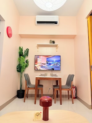 Ein modernes Zimmer mit Esstisch, zwei Stühlen, Fernseher, Klimaanlage, einer Uhr, einem Kaktus und einem Feuerlöscher.