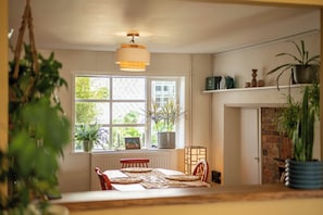 A cozy room with a dining table, chairs, a hanging light fixture, a window with plants, and a brick fireplace. - Cosy Sussex Cottage (Robertsbridge, Staplecross)