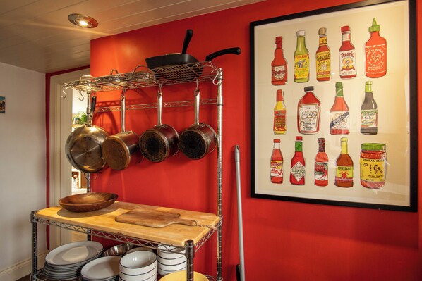 A kitchen with pots and pans hanging on a rack, a wooden table with dishes, and a wall with a framed illustration of condiments.