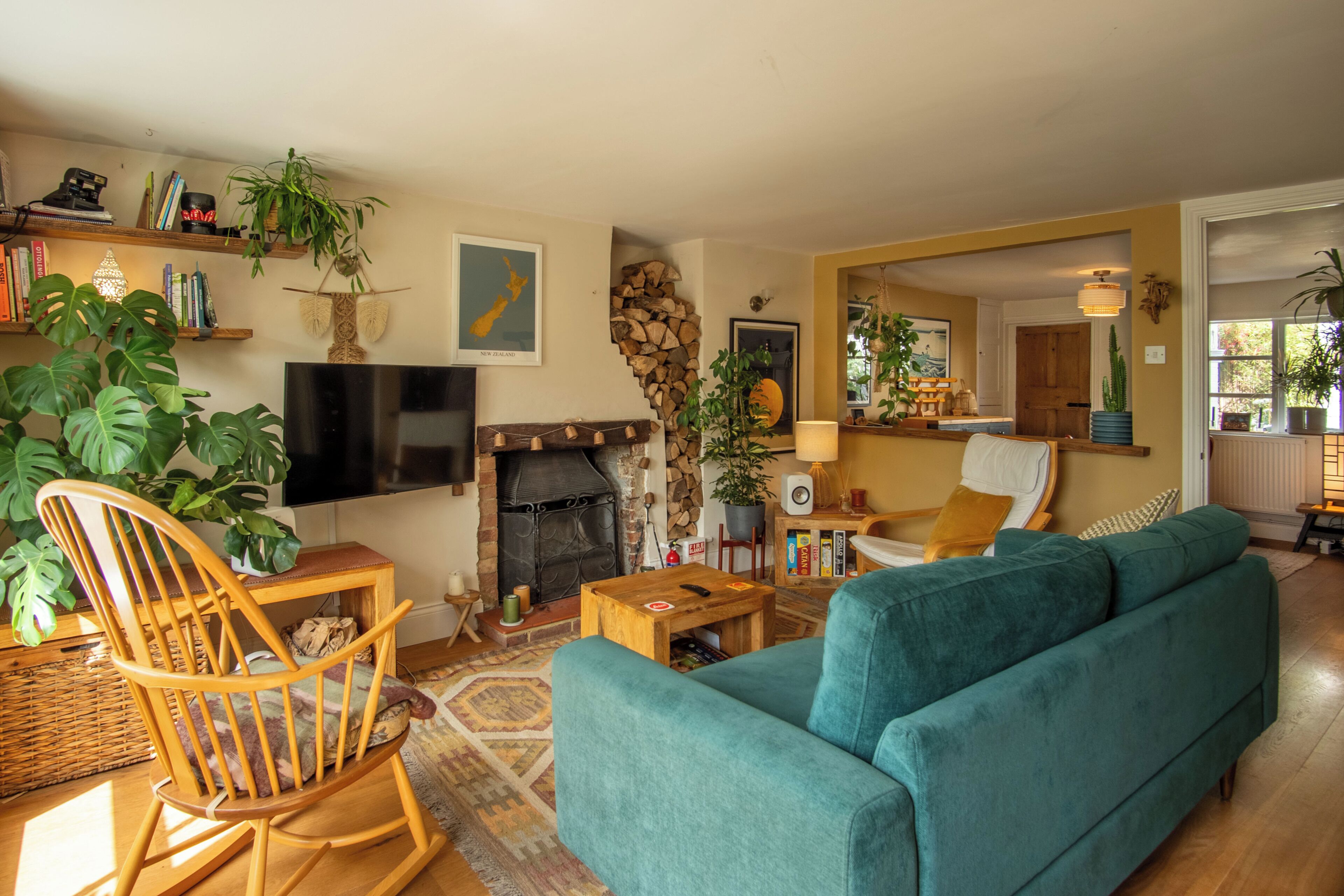 A cozy living room with a fireplace, a teal sofa, wooden rocking chair, and various plants.