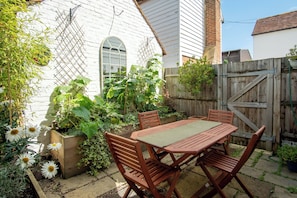 A wooden table and chairs set in a garden with a white brick wall, wooden fence, and potted plants. - Cosy Sussex Cottage (Robertsbridge, Staplecross)