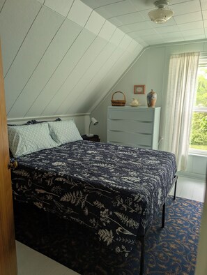 Ein Schlafzimmer mit schrägen Wänden, einem Bett mit blumigem Überwurf, einer Kommode, einer Lampe und einem Fenster mit Vorhängen.