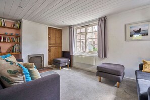 A cozy living room with a fireplace, a bookshelf, a window with curtains, and a framed picture on the wall. - Host & Stay | Teal Cottage (Burnham Overy Staithe)