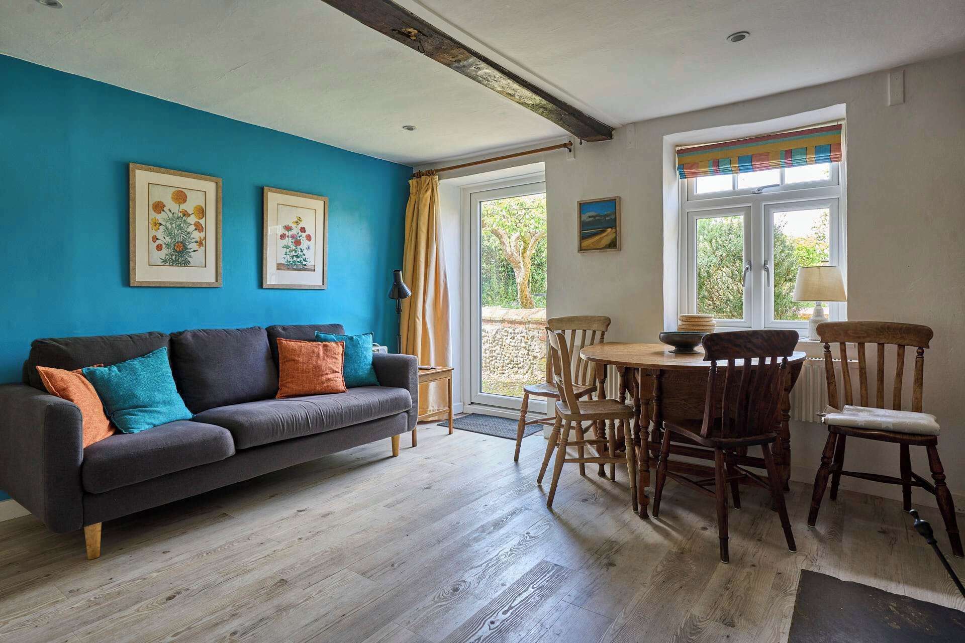 A room with a blue wall, a grey sofa with colorful cushions, a wooden dining table with chairs, and a window with a view of greenery.