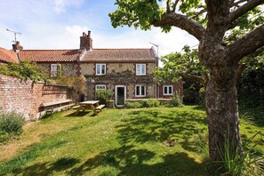 A two-story stone house with a tiled roof, a front door, and a small front yard with a bench. - Host & Stay | Teal Cottage (Burnham Overy Staithe)