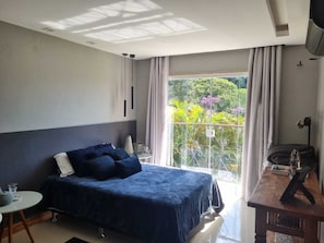 Ein Schlafzimmer mit Bett, einem kleinen Tisch, einem Stuhl, einem Fernseher und einem Balkon mit Blick auf Grünfläche.