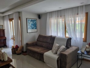 A living room with a brown sofa, a framed artwork on the wall, and a window with sheer curtains. - Casa Azaléia  (Teresópolis)