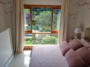 A bedroom with a large window, a bed with pink bedding, and a view of a garden. - Casa Azaléia  (Teresópolis)