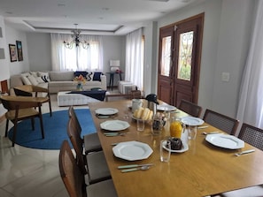 Ein Essbereich mit einem langen Tisch, gedeckt für vier Personen, umgeben von Stühlen. Im Hintergrund befindet sich eine Couch und ein kleiner Tisch mit einer Lampe.