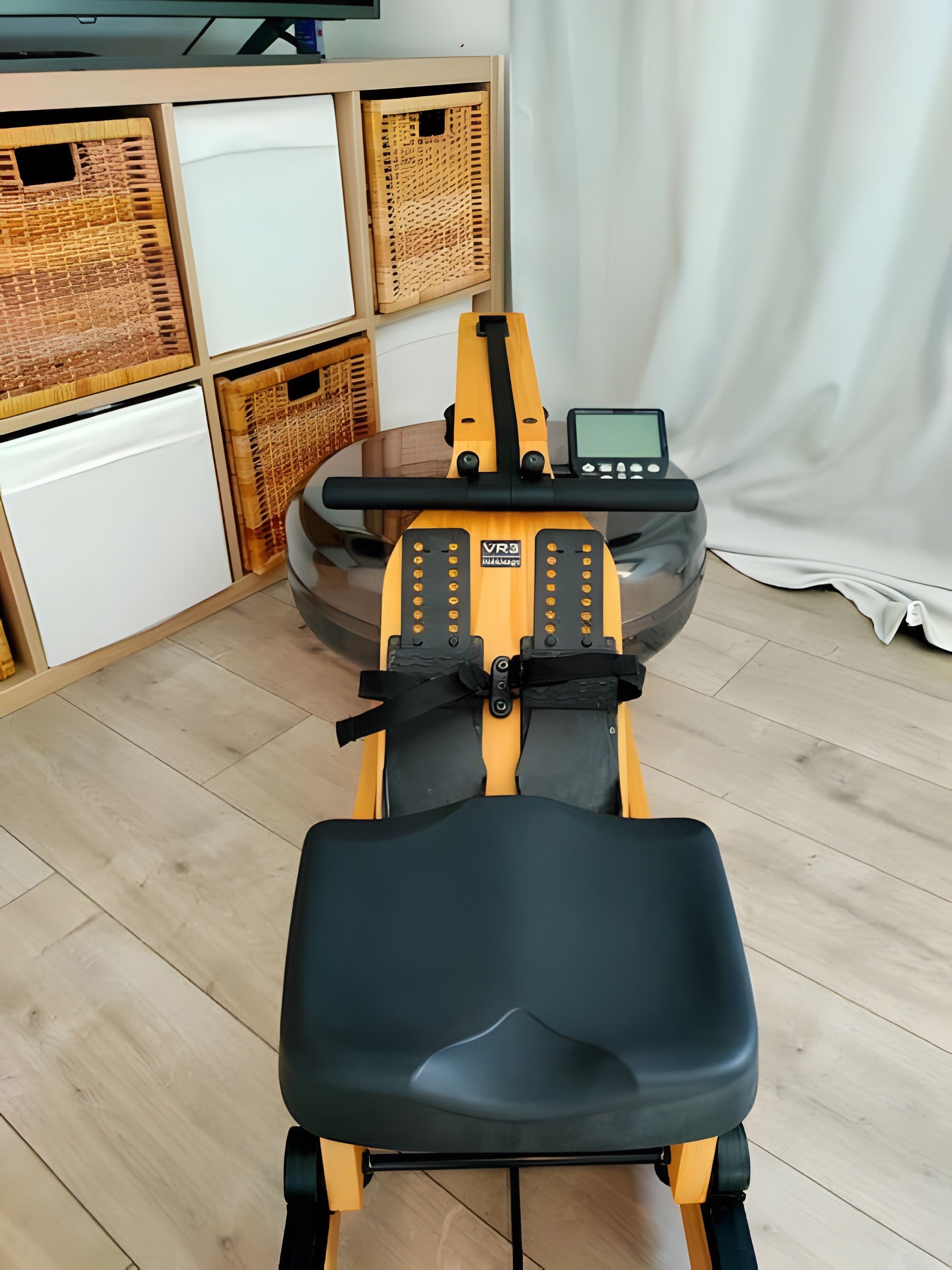 Un vélo d’appartement, doté d’un écran numérique, est placé dans une pièce dont le sol est recouvert de parquet et qui contient des paniers en osier.