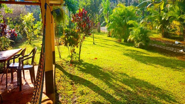 Uma área externa coberta com mesa e cadeiras, cercada por vegetação exuberante e árvores.