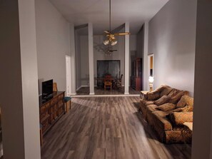 Ein geräumiges Wohnzimmer mit Holzboden, einer Couch, einem Fernseher und einem Deckenventilator.