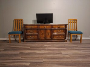 Eine hölzerne Kommode mit Schubladen und einem Fernseher darauf, begleitet von zwei Holzstühlen mit blauen Kissen.