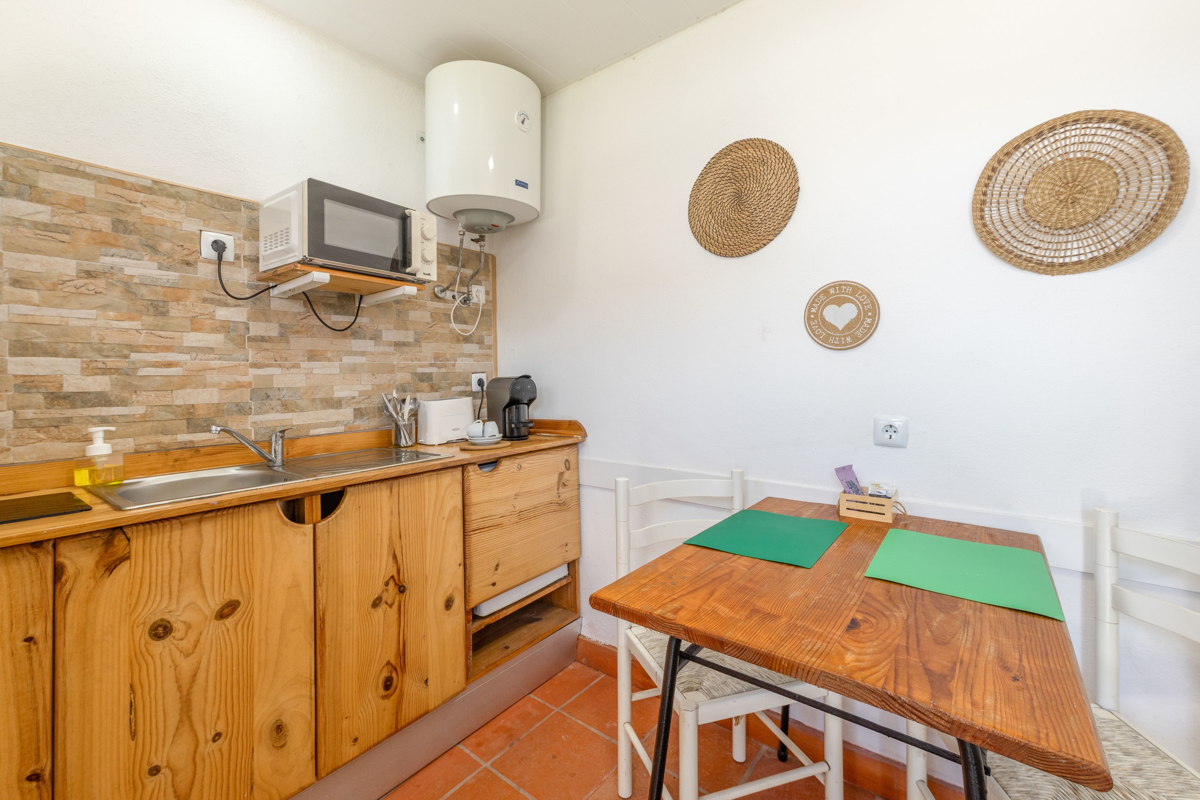 A compact kitchen with a microwave, kettle, and coffee maker, a wooden table with a green placemat, and two woven wall decorations.