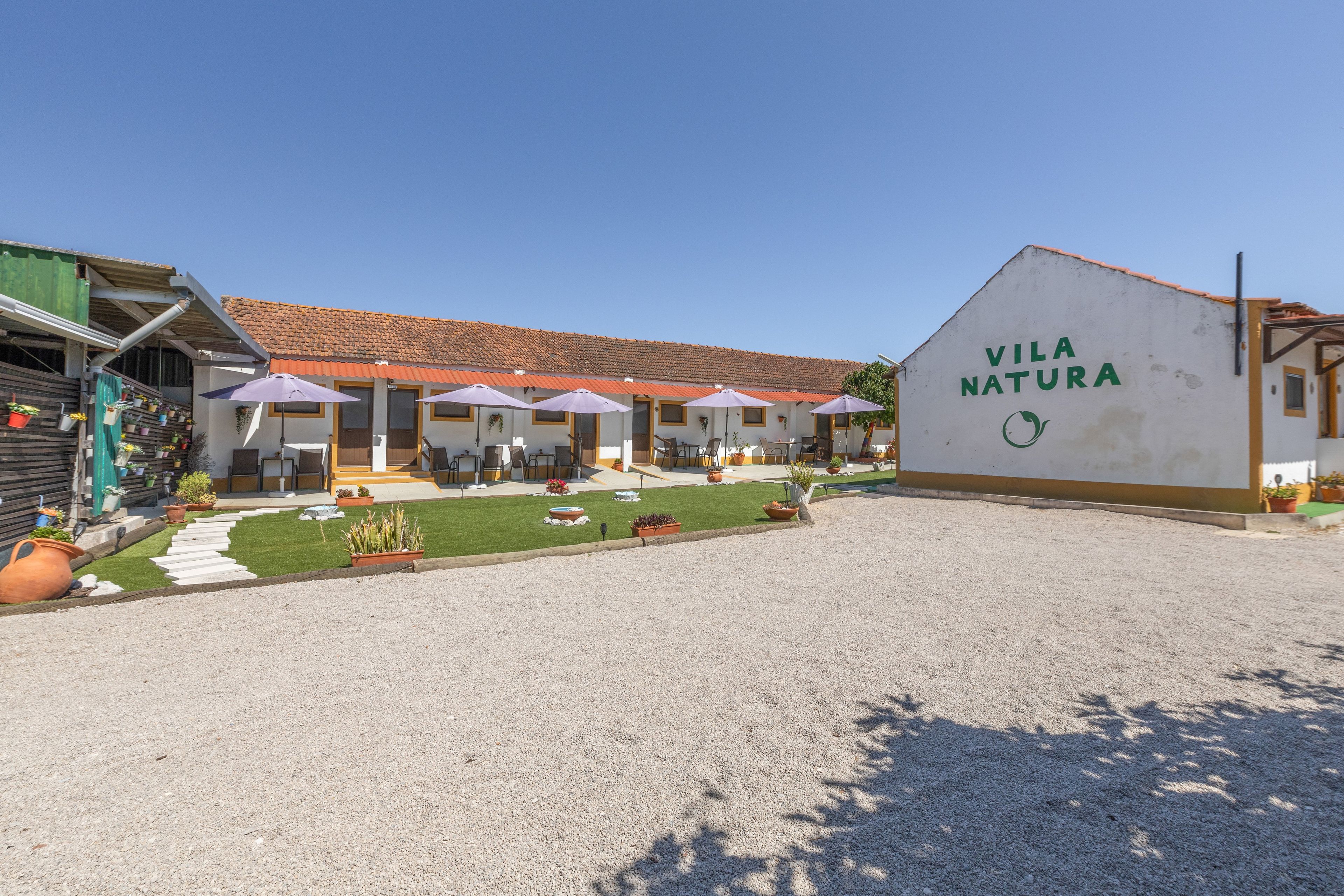 Un bâtiment sur lequel on peut lire l’inscription « VILA NATURA », et une zone de sièges extérieure abritée par un auvent et dotée de parasols.