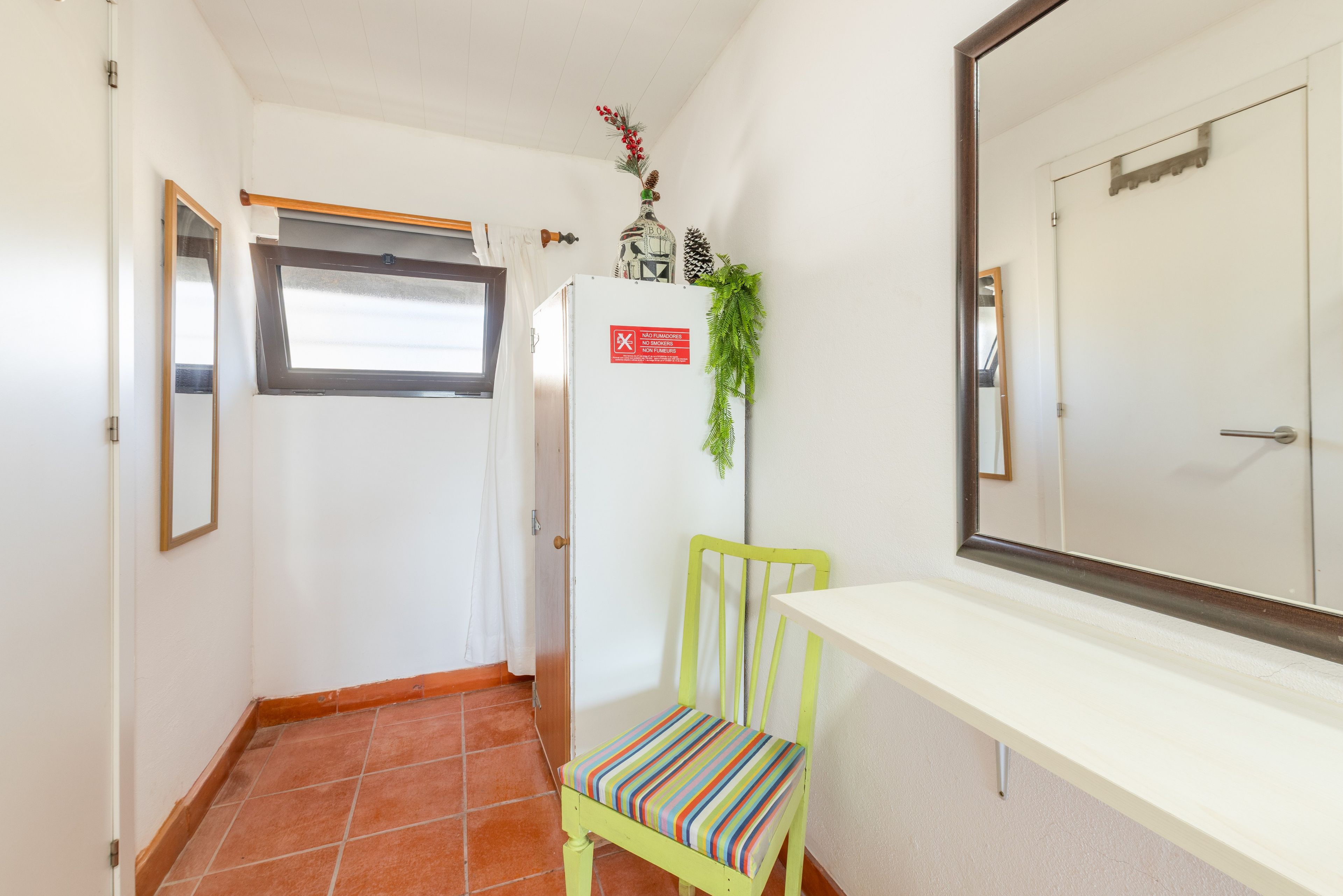 A narrow hallway with a window, a refrigerator, a chair, and a mirror.