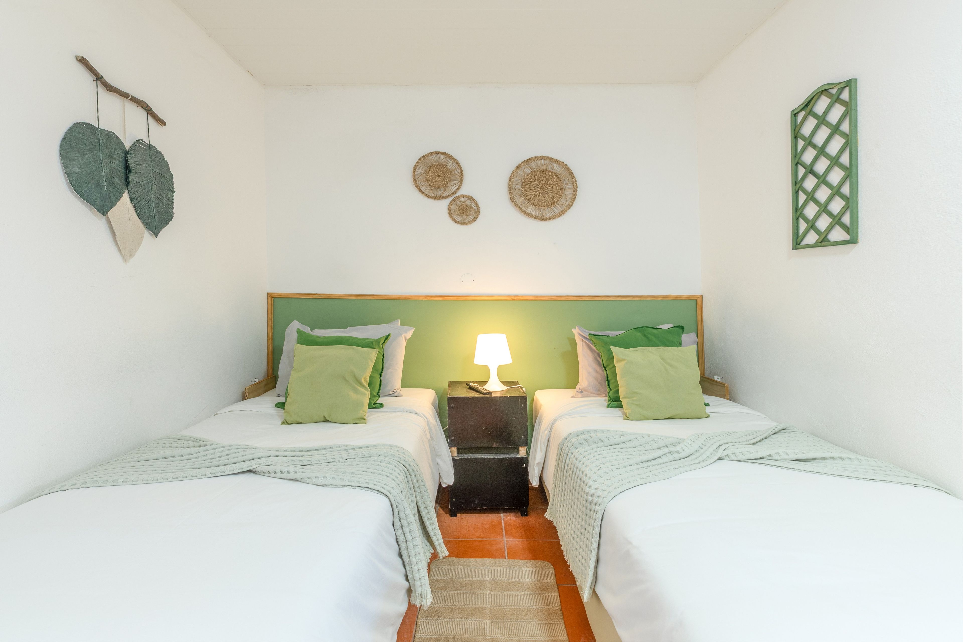 Una habitación con dos camas, una mesita de noche con lámpara y adornos en la pared, incluyendo un adorno colgante con forma de corazón y elementos tejidos circulares.