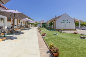 Pátio com área de estar externa, gramado verde e um edifício com a inscrição 'Vila Natura'.