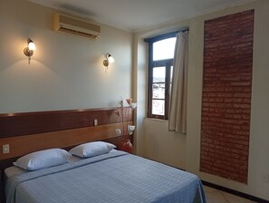 Ein Schlafzimmer mit einem hölzernen Kopfende, einem Bett mit blauer Bettwäsche, zwei Wandleuchten, einem Fenster mit Vorhängen und einer Ziegelwand.