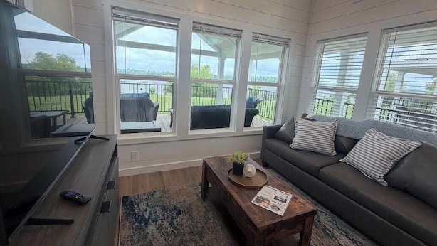 Ein Wohnzimmer mit einer Couch, einem Couchtisch und einem Fernseher. Es gibt Fenster mit Jalousien und einen Blick ins Freie.