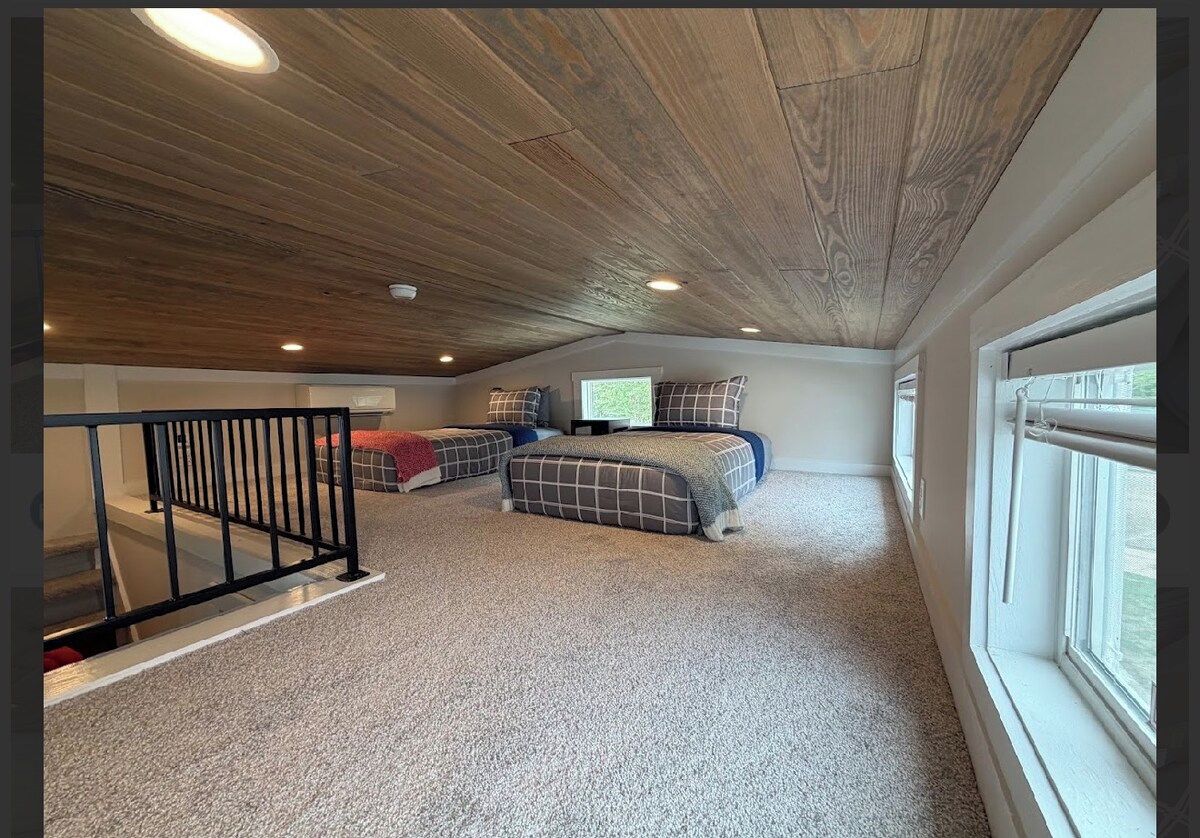 Una habitación con techo inclinado, una cama con colcha a cuadros, una ventana y una escalera.