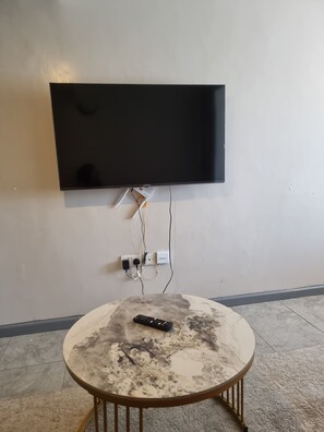 Ein Flachbildfernseher an der Wand, eine Fernbedienung auf einem runden Tisch und eine Mehrfachsteckdose mit Kabeln.