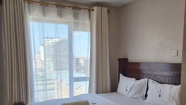 A hotel room with a bed, curtains, a window with a view of buildings, and a light fixture. - Modern Studio in Fedha, Nairobi (Nairobi)