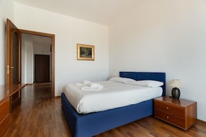 Un dormitorio con una cama, una mesita de noche, una lámpara, un cuadro enmarcado en la pared y suelo de madera.