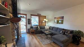 A living room with a grey sofa, wooden flooring, a chessboard, and a view of a house through the window. - Modern Mountain Retreat - Walk to Village + HotTub (Beaverdell)