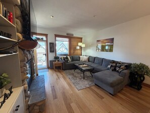 Ein Wohnzimmer mit grauem Sofa, Holzboden, einem Schachbrett und Blick auf ein Haus durch das Fenster.