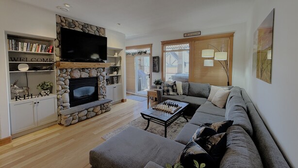 A living room with a stone fireplace, a sofa, a coffee table, and a TV. - Modern Mountain Retreat - Walk to Village + HotTub (Beaverdell)