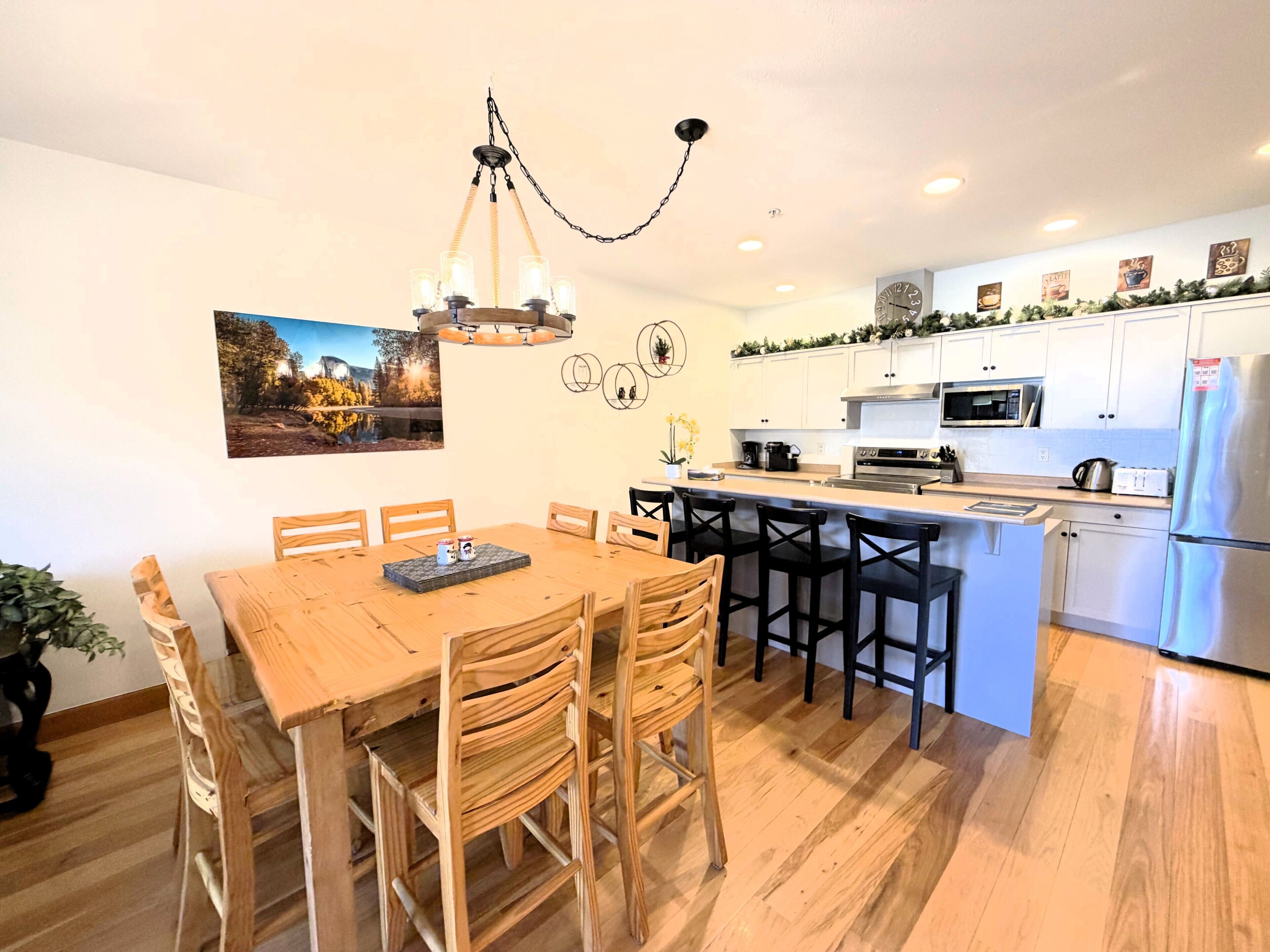 A modern kitchen with wooden dining table and chairs, a built-in island with bar stools, stainless steel appliances, and a decorative light fixture.