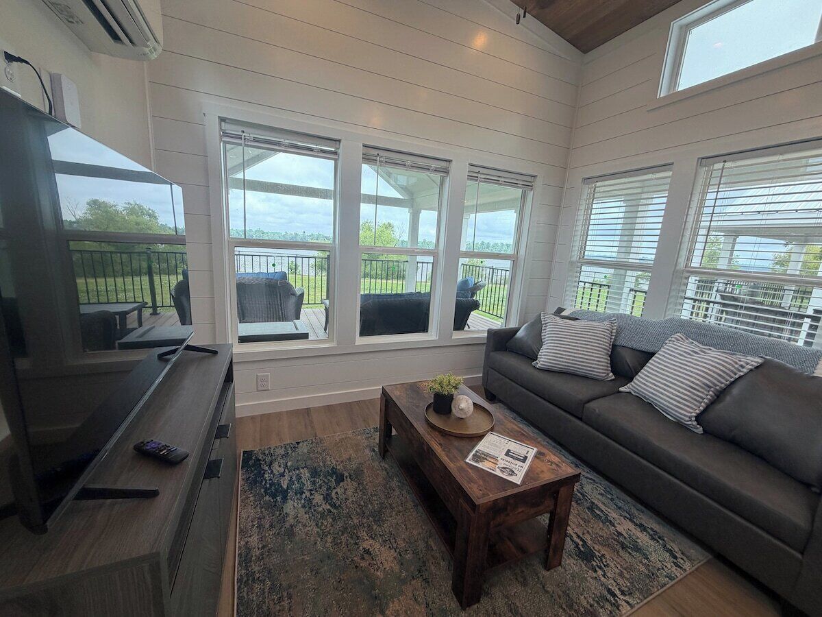 A living room with a sofa, coffee table, and TV. There are large windows with blinds, a balcony, and a view of greenery.