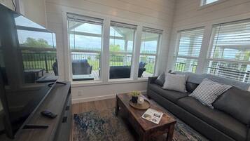A living room with a sofa, coffee table, and TV. There are large windows with blinds, a balcony, and a view of greenery.