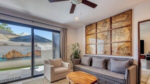 A modern living room with a sofa, armchair, and coffee table, a large window with a view of a rocky landscape, and a wall-mounted map. - Family-friendly Oasis With Pool and Steps From Medano Beach (Cabo San Lucas)