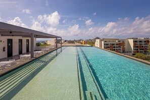 Una piscina con agua cristalina, un área de descanso techada y edificios al fondo.