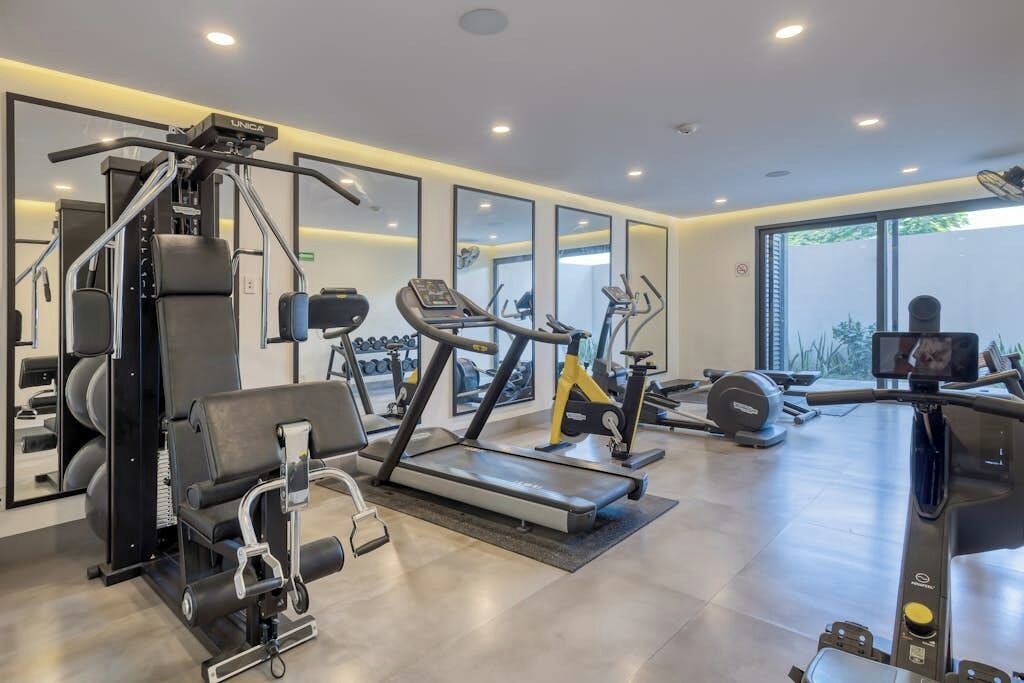 Une salle de sport moderne équipée de divers appareils d’exercice, de grands miroirs et offrant une vue sur l’extérieur.