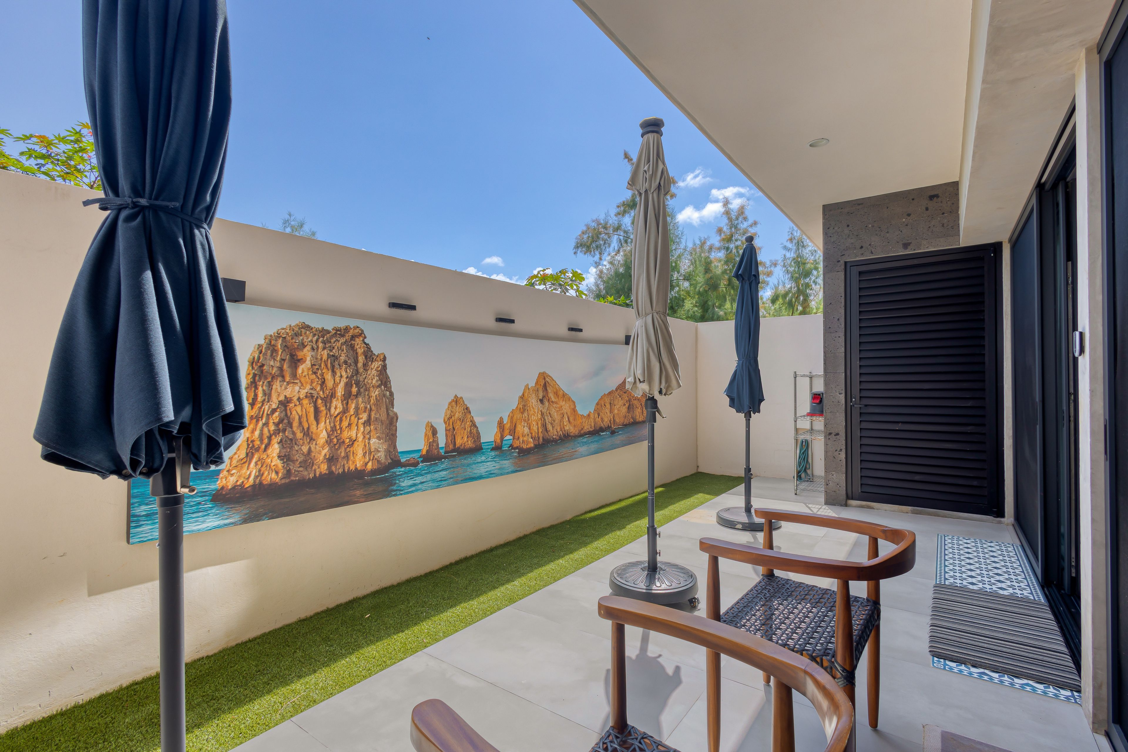 Zona d'estar exterior moderna amb un mural d'una escena costanera, una ombrell terrassa i una porta que dona a l'interior.