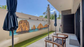 Zona d'estar exterior moderna amb un mural d'una escena costanera, una ombrell terrassa i una porta que dona a l'interior.