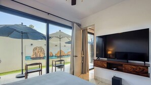 A modern hotel room with a large bed, a TV mounted on the wall, and a view of a pool and rocks through the glass door. - Family-friendly Oasis With Pool and Steps From Medano Beach (Cabo San Lucas)