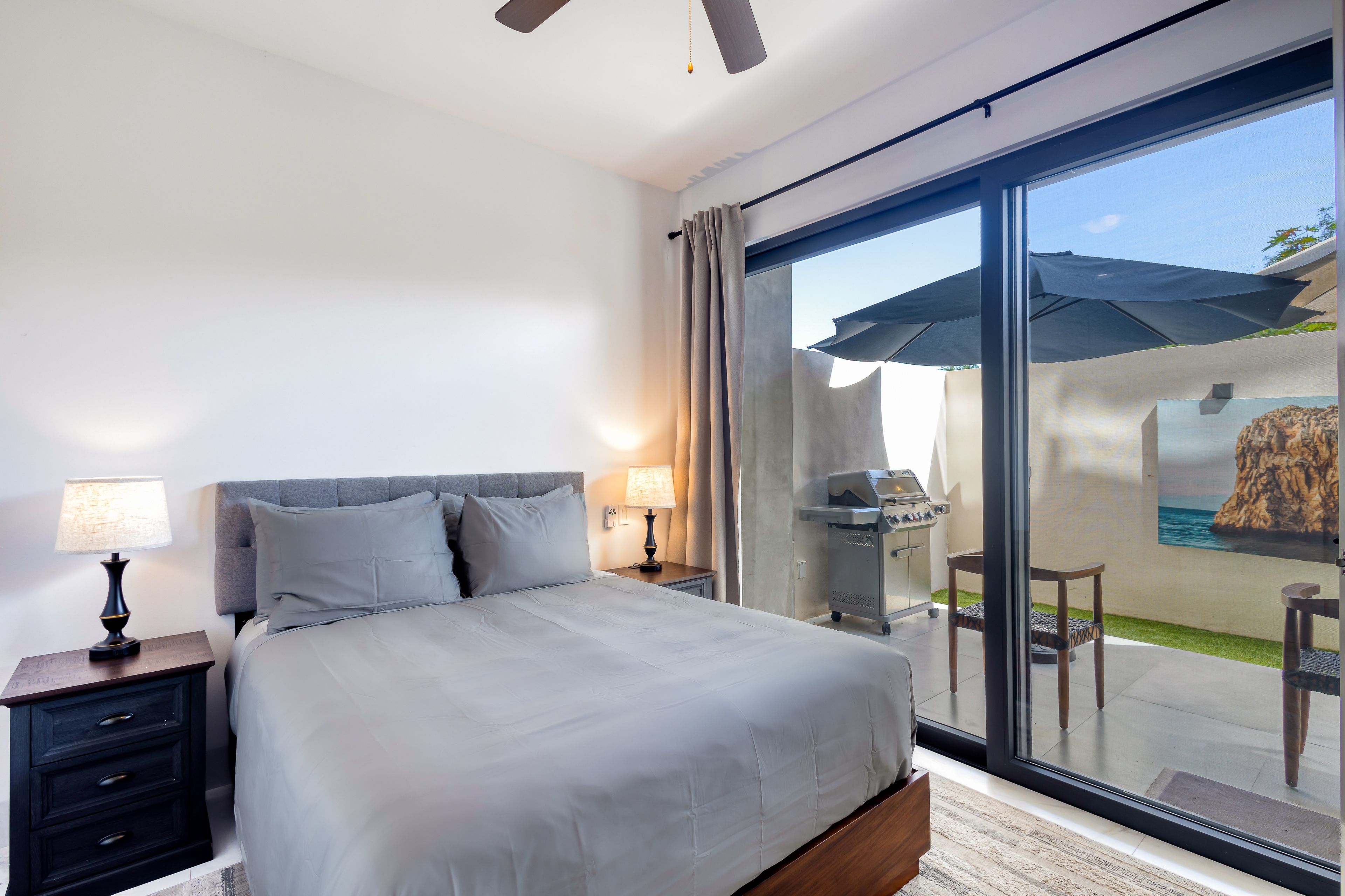 A modern bedroom with a large bed, bedside lamps, a nightstand, and a sliding glass door leading to an outdoor area with a table and chairs.