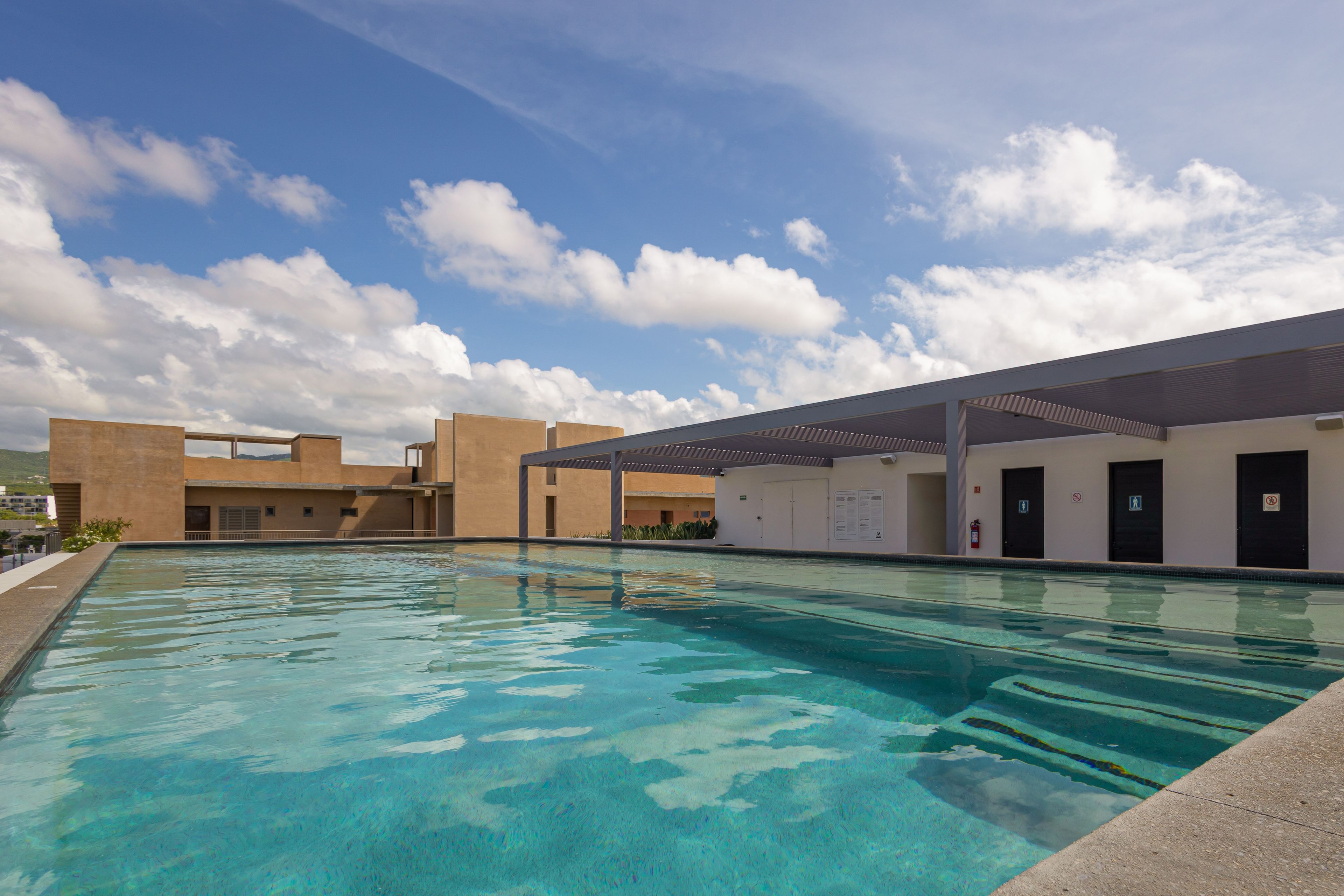 A swimming pool with clear blue water, surrounded by a building with multiple doors and windows, under a partly cloudy sky.