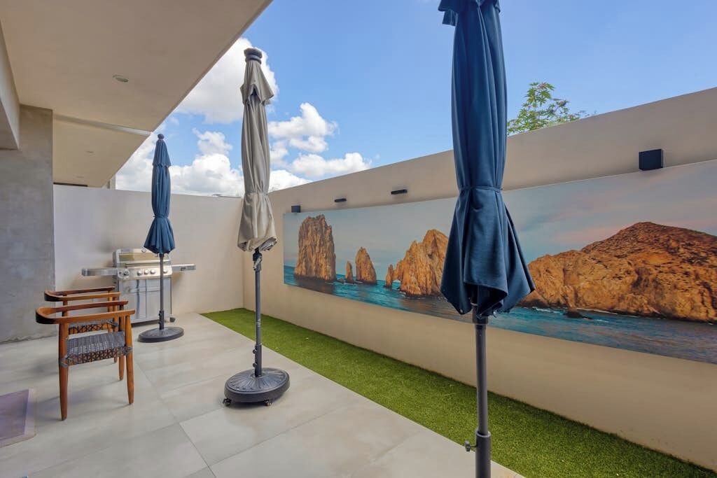 An outdoor patio area with a grill, umbrellas, a chair, and a mural of a coastal landscape.