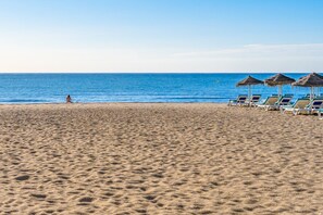 Ein Sandstrand mit klarem, blauem Meer, eine Person sitzt im Sand und es gibt mehrere Liegestühle mit Strohdächern.