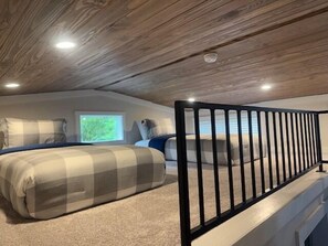 Un dormitorio con techo inclinado, una cama con manta de cuadros, un ventanal y una barandilla negra.