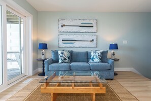 A living room with a blue sofa, two lamps, a glass coffee table, and two wall-mounted paintings. - Bay Breeze Retreat: Lakefront Condo with Pool (Put-in-Bay)