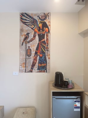 A wall-mounted artwork of an Egyptian figure, a small kitchenette with a coffee maker, and a refrigerator.