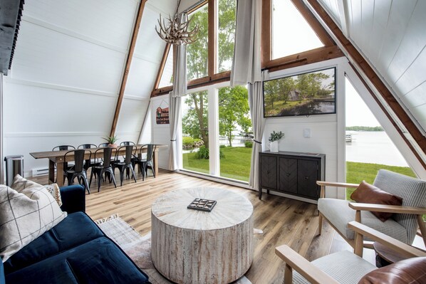 A spacious living area with a round coffee table, a sofa, and a dining table with chairs. Large windows offer a view of a lake and greenery.